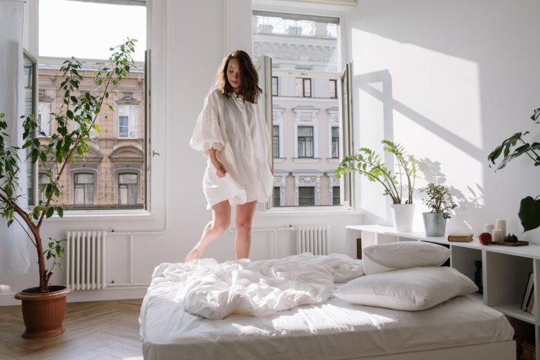 A woman enjoys a peaceful morning in a sunlit bedroom with open windows and potted plants.