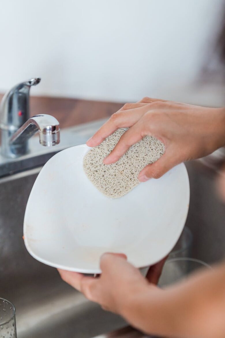 A person scrubs a white plate using a sponge under a kitchen faucet indoors.