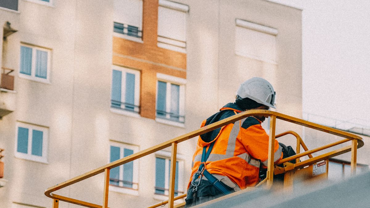 A construction worker in safety gear operates a lift near an apartment building. A construction worker in safety gear operates a lift near an apartment building.