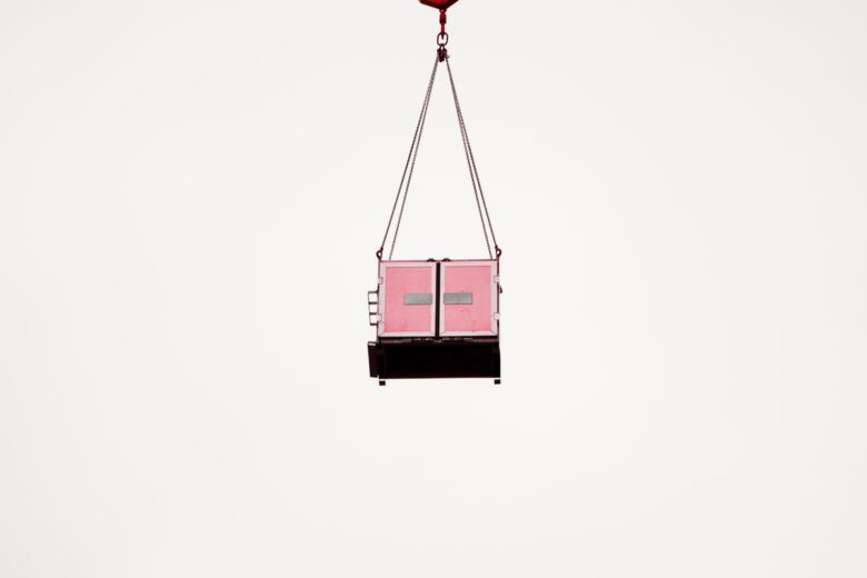 A pink cargo container suspended mid-air against a clear white sky, minimalist composition.