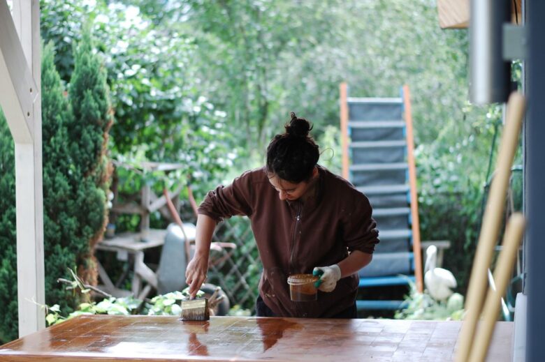 Woman applies varnish to a table outdoors, surrounded by greenery. DIY and home improvement scene.