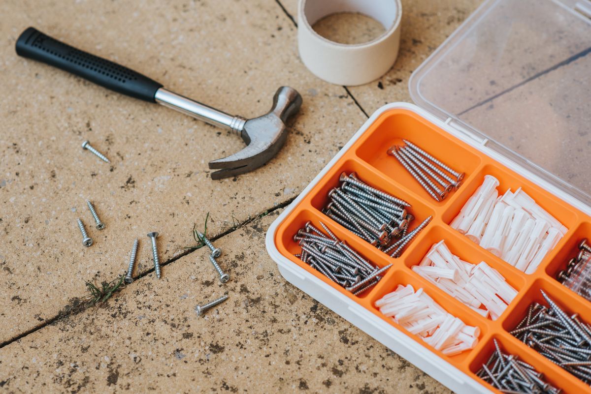 Top view of assorted repair instruments and screws in box near hammer and scotch tape on dirty tile in soft daylight Top view of assorted repair instruments and screws in box near hammer and scotch tape on dirty tile in soft daylight