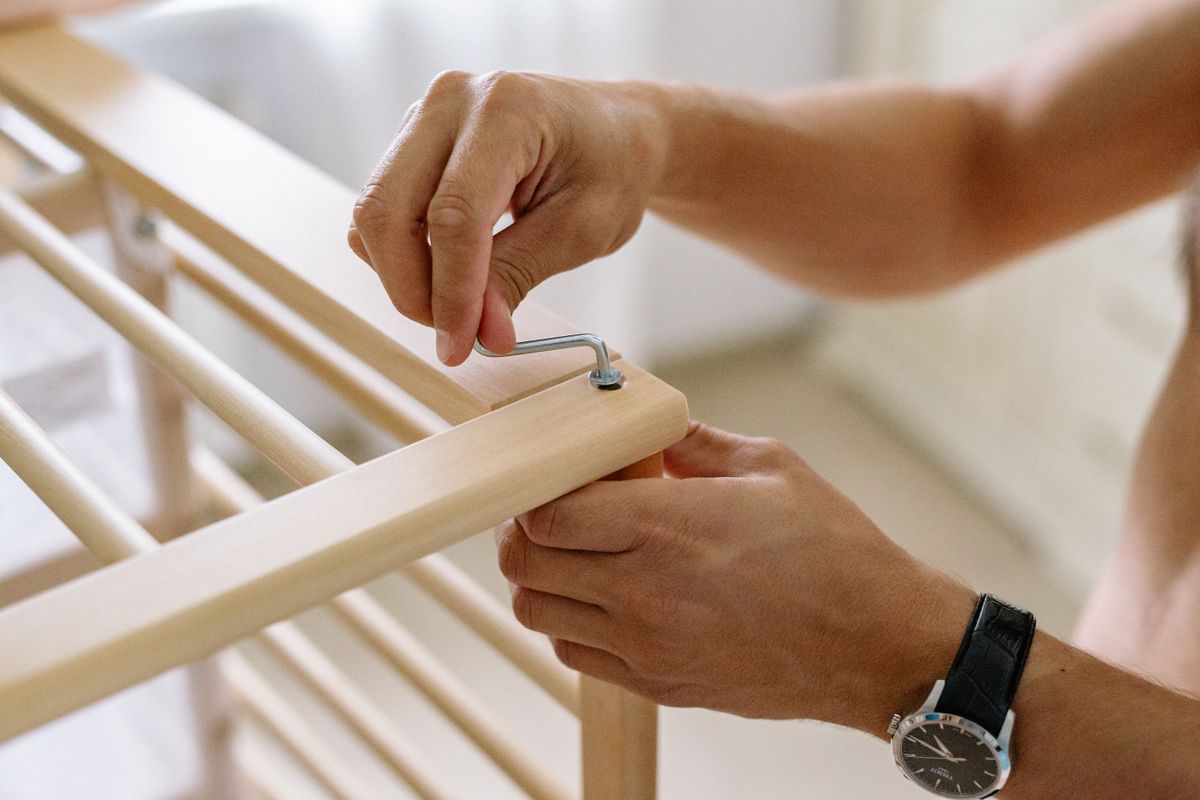 Close-up of a man using an Allen wrench to assemble a wooden baby cot indoors. Close-up of a man using an Allen wrench to assemble a wooden baby cot indoors.