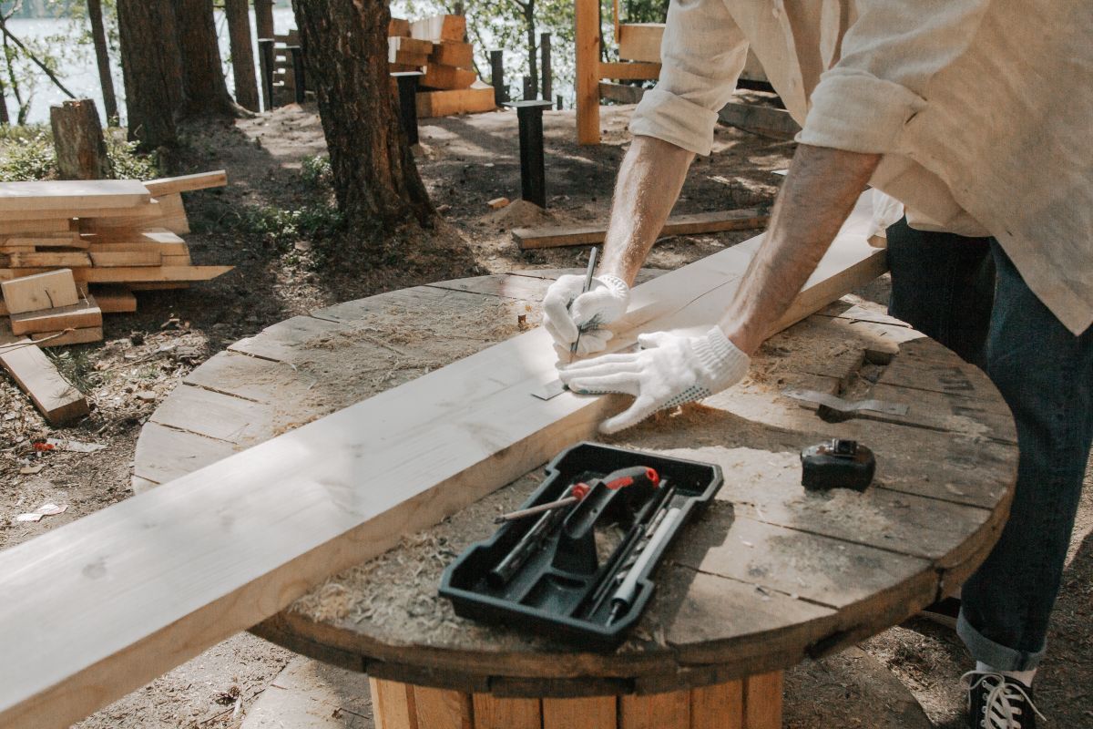 A carpenter skillfully outlines on a wooden plank with tools outdoors, surrounded by trees. A carpenter skillfully outlines on a wooden plank with tools outdoors, surrounded by trees.