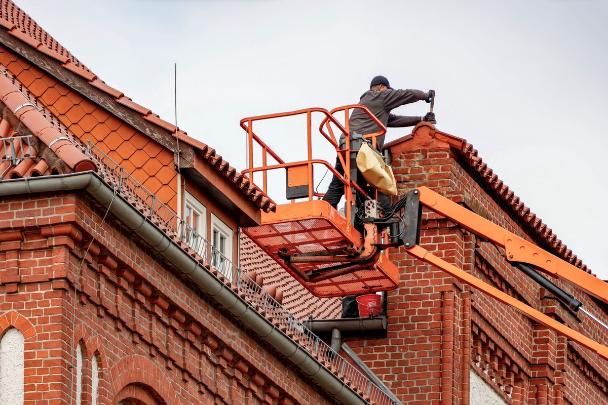 Roofer in a cherry picker conducting maintenance on a red brick building with a tiled roof. Roofer in a cherry picker conducting maintenance on a red brick building with a tiled roof.
