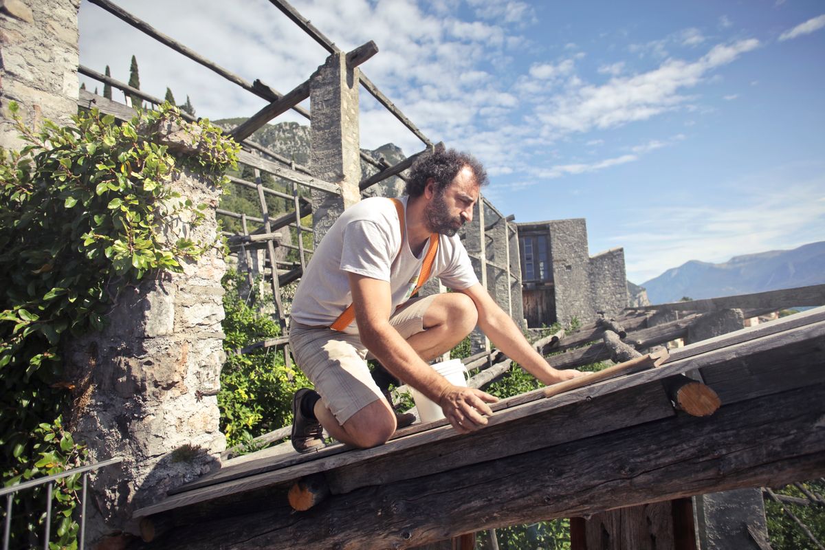 Side view of male builder in summer wear reconstructing rooftop and sitting on wooden beams of aged building on background of scenic landscape in sunny day Side view of male builder in summer wear reconstructing rooftop and sitting on wooden beams of aged building on background of scenic landscape in sunny day