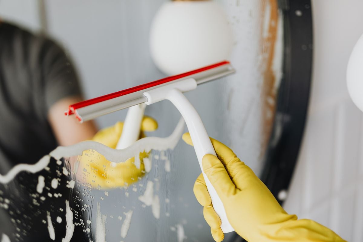 Faceless person in yellow rubber gloves using scraper to remove soapy water from mirror in bathroom Faceless person in yellow rubber gloves using scraper to remove soapy water from mirror in bathroom