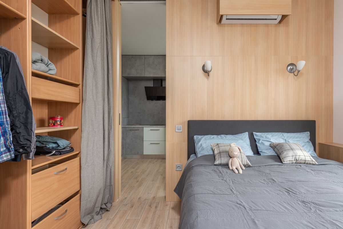 Modern room interior with bed with pillows and soft rabbit toy near wardrobe with shelves and clothes near entrance to kitchen with curtains in light apartment