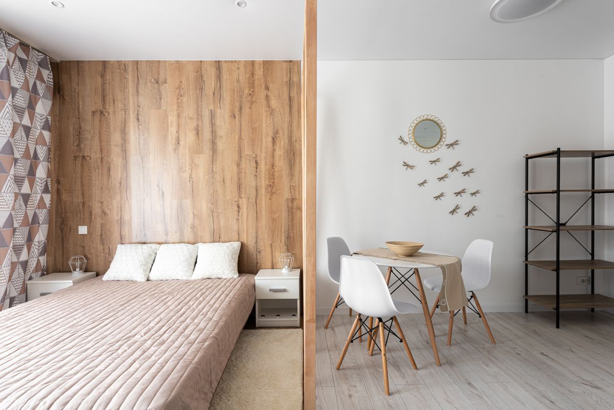 Bed with cushions against table and chairs on floor at home with empty shelves and decorative dragonflies on wall