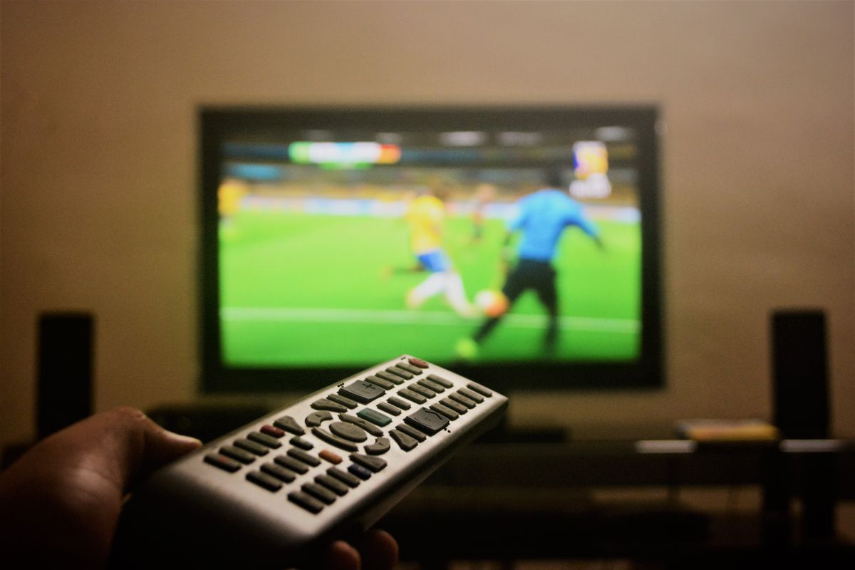 Close-up of a hand holding a remote control in front of a TV showing a football match. Close-up of a hand holding a remote control in front of a TV showing a football match.
