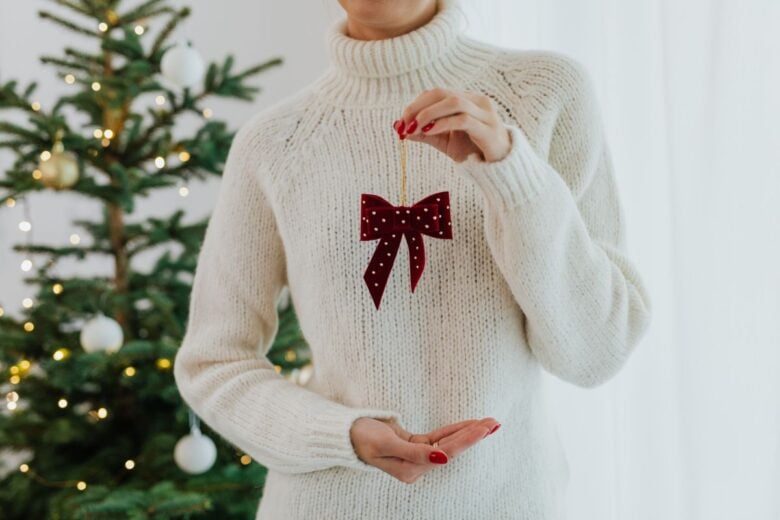 A person holds a red bow ornament by a Christmas tree in a cozy white sweater.