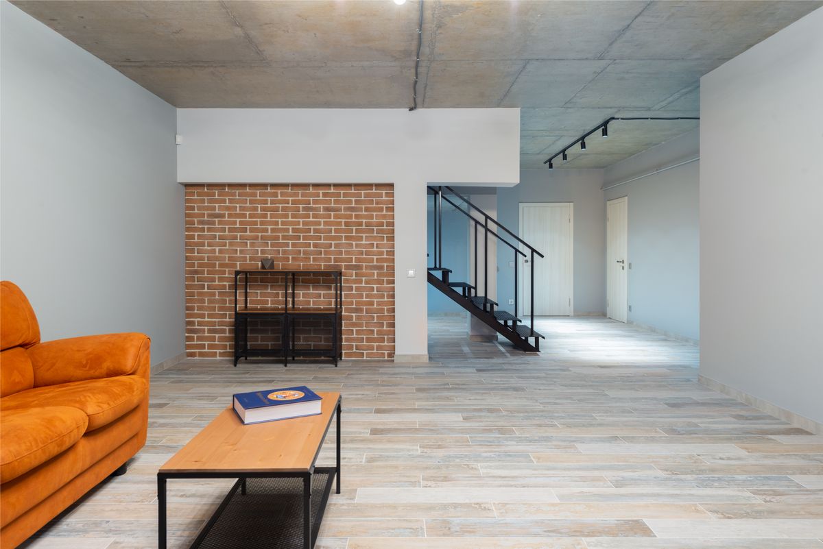 Orange comfortable sofa near table with book placed in stylish light spacious apartment with stairway brick wall and closed doors